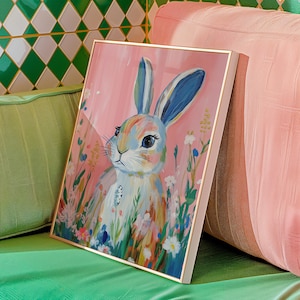 May include: A framed painting of a colorful rabbit in a field of flowers. The rabbit is painted in shades of blue, pink, and green, and the flowers are a mix of pink, white, and yellow. The background is a soft pink.