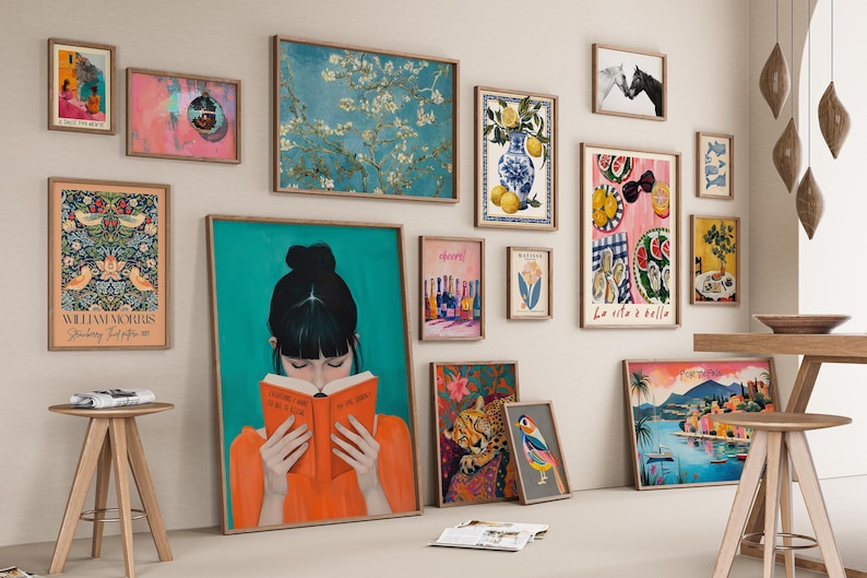 May include: A gallery wall with a variety of framed artwork, including a painting of a woman reading a book, a floral print, a still life with lemons and a vase, and a landscape scene. The wall is painted a light beige color and the frames are all in a natural wood finish.