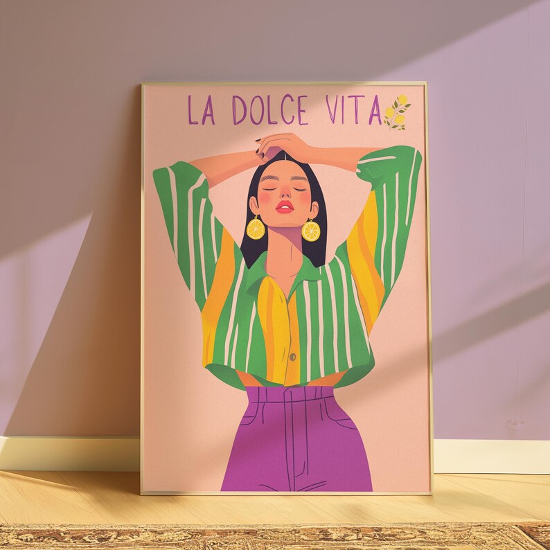 May include: A framed art print featuring a woman with her arms raised, wearing a striped green and yellow shirt, purple pants, and lemon earrings. The text "LA DOLCE VITA" is at the top. The artwork has a soft, colorful, and illustrative style.