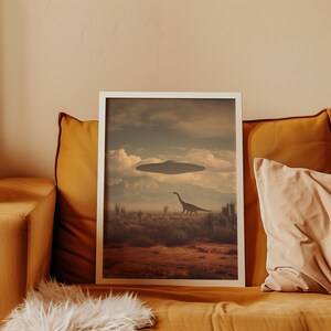 May include: A framed art print featuring a surreal landscape with a dinosaur and a UFO hovering in the sky. The artwork has a vintage aesthetic with a color palette of browns, tans, and blues. The print is displayed on a mustard-colored couch.
