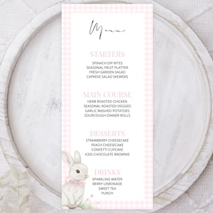 May include: A menu card with a pink gingham border and a watercolor illustration of a bunny wearing a pink bow. The menu lists starters, main courses, desserts, and drinks. The menu is on a white wooden plate.