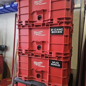 May include: Red Milwaukee Packout storage boxes stacked. The boxes have the Milwaukee logo and the words "PACKOUT" printed in white. Two boxes have labels that read "2-GANG BOXES" and "MUD RINGS".