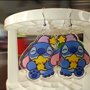 May include: A pair of clear acrylic earrings featuring a cartoon character with blue fur, pink ears, and a purple hat. The character is holding a yellow star. The earrings have silver-colored fish hook ear wires.