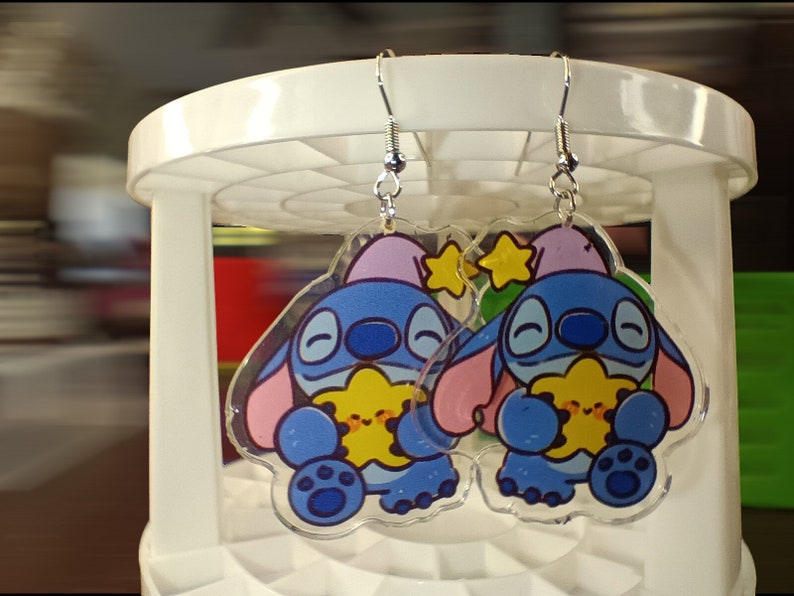 May include: A pair of clear acrylic earrings featuring a cartoon character with blue fur, pink ears, and a purple hat. The character is holding a yellow star. The earrings have silver-colored fish hook ear wires.