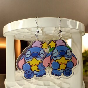 May include: A pair of clear acrylic earrings featuring a cartoon character in blue, pink, and yellow. The earrings have silver-colored fish hook ear wires. The character is holding a yellow star.