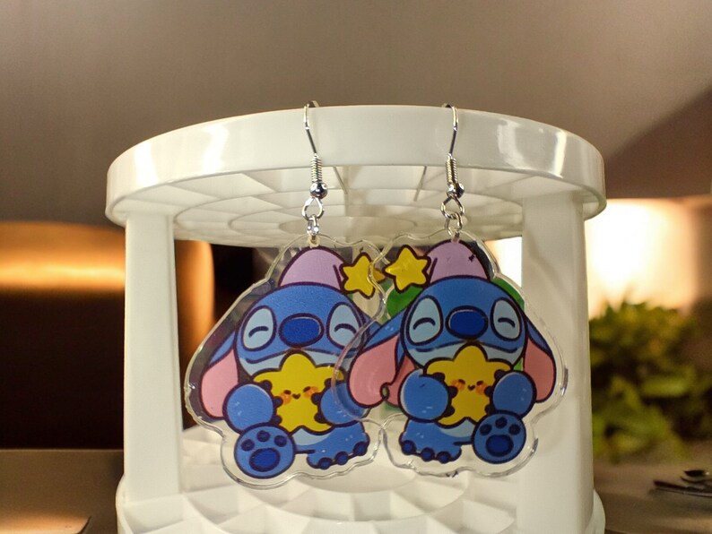 May include: A pair of clear acrylic earrings featuring a cartoon character in blue, pink, and yellow. The earrings have silver-colored fish hook ear wires. The character is holding a yellow star.