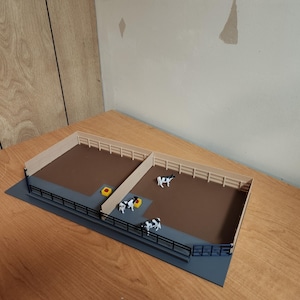 Peut inclure: Une scène de ferme miniature avec deux enclos clôturés, un sol brun, des clôtures beiges et des garde-corps noirs. Plusieurs vaches jouets noires et blanches sont placées à l'intérieur des enclos. Une mangeoire jaune est également présente.