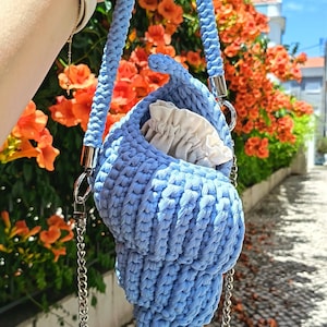 May include: A light blue crochet handbag shaped like a seashell, featuring a thick braided handle, a silver-toned metal chain strap, and a cream-colored fabric drawstring closure tucked inside the main compartment of the bag.
