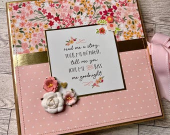 May include: A handmade scrapbook with a floral design and the text "read me a story, tuck me in tight, tell me you love me and kiss me goodnight." The cover features pink and gold accents, with decorative flowers and a pink ribbon.