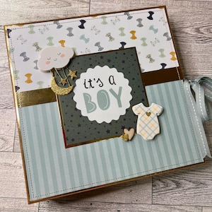 May include: A baby scrapbook album with a blue and gold theme. The cover features the text "it's a BOY" surrounded by decorative elements like a cloud, stars, and a baby onesie. The album is bound with a light blue ribbon.