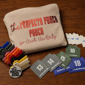 May include: A beige fabric pouch with the red text "the PERFECTA PUNCH POUCH for Cinch Use Only" printed on the front. Included are a set of multi-colored plastic punch guides on a chain, a white circular tag, and several numbered white, gray, green, and blue plastic tags.