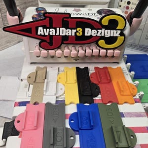 May include: A collection of colorful, 3D-printed tools, including various colored guides and a central device with the text "AvaJDar3 Dezignz." The guides are in shades of beige, yellow, black, red, orange, pink, gray, blue, and green.
