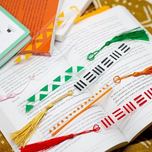 Op de afbeelding: Een set van vijf transparante acryl bladwijzers met verschillende gekleurde kwastjes en geometrische ontwerpen. De bladwijzers hebben zwarte, rode, oranje, groene en gele accenten.