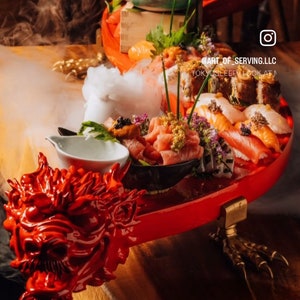 Puede incluir: Un plato de servir rojo en forma de dragón con detalles dorados, lleno de una variedad de sushi, sashimi y otros platos japoneses. El plato está sobre una mesa de madera con un fondo humeante. El texto "@ART_OF_SERVING.LLC" y "TOKYOSLEEPER LOOK AT ME" es visible en el plato.