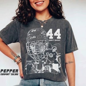 Op de afbeelding: Donkergrijs T-shirt met een afbeelding van een hockeyspeler, het nummer 44 en de naam Garrett Graham. Het shirt heeft een vintage uitstraling en de woorden "Pepper Comfort Colors" staan onderaan gedrukt.