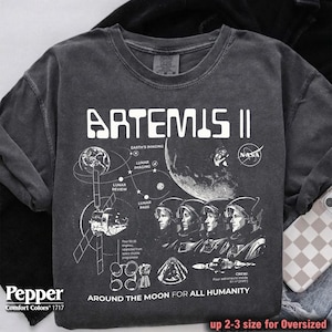Puede incluir: Camiseta gráfica gris oscuro de Artemis II con el logotipo de la NASA. El diseño incluye ilustraciones de la luna, astronautas y diagramas técnicos. El texto dice "AROUND THE MOON FOR ALL HUMANITY".
