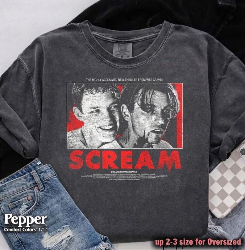 May include: Dark gray sweatshirt featuring a graphic from the movie "Scream." The design includes two faces, the title "SCREAM" in red, and text that reads "THE HIGHLY ACCLAIMED NEW THRILLER FROM WES CRAVEN." The sweatshirt is paired with denim shorts.