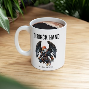 Puede incluir: Taza de cerámica blanca con una ilustración en negro y naranja de un pájaro con un casco y el texto "Derrick Hand" y "Rack 'em & Stack 'em".