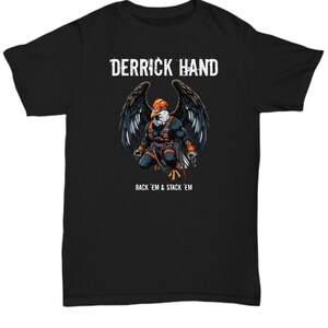 Puede incluir: Camiseta negra con un gráfico de un águila calva con un casco y sosteniendo una cuerda. El águila está posada en una viga de madera. El texto "Derrick Hand" y "Rack 'em & Stack 'em" está impreso en la camiseta.