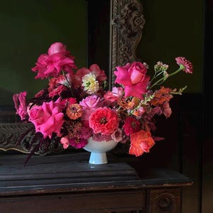 Op de afbeelding: Een levendig bloemstuk in een witte voetbakschaal. Het boeket bevat een verscheidenheid aan roze en rode bloemen, waaronder rozen en zinnia's, met vleugjes oranje en perzik. De opstelling staat op een donkere houten ondergrond.