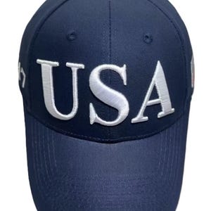 May include: Navy blue baseball cap with the word "USA" in white, bold, raised lettering across the front. The cap has a curved brim and a button on top. The cap is a solid color with a structured fit.