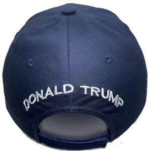 May include: A navy blue baseball cap with the embroidered text "DONALD TRUMP" in white across the back. The cap has a curved brim and a button on top.