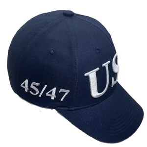 May include: A navy blue baseball cap with the white letters "US" embroidered on the front. The number "45/47" is embroidered in white on the side. The cap has a curved brim and a button on top.