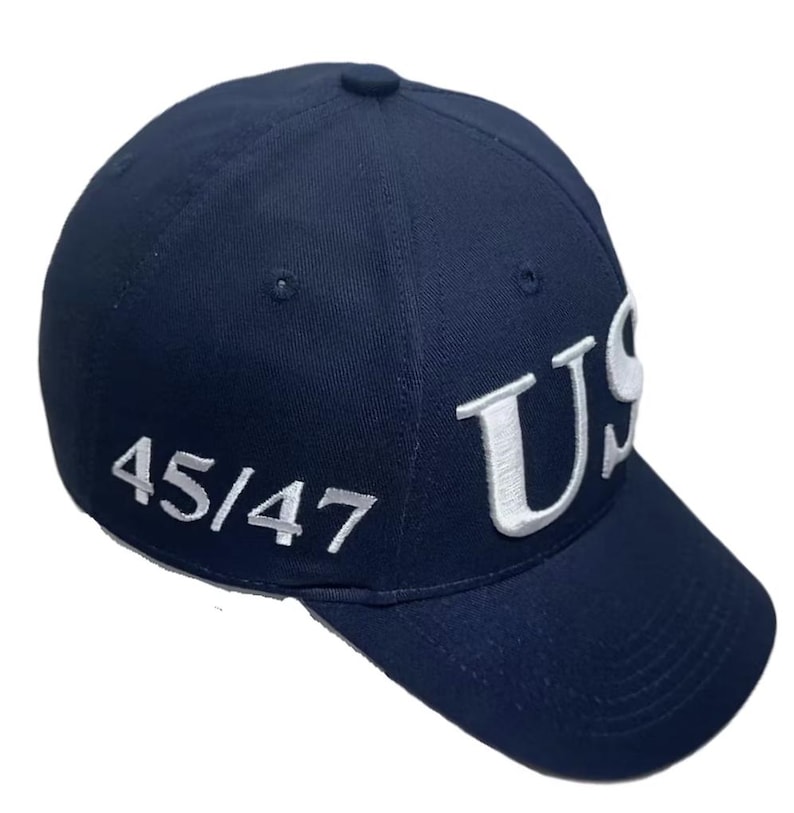 May include: A navy blue baseball cap with the white letters "US" embroidered on the front. The number "45/47" is embroidered in white on the side. The cap has a curved brim and a button on top.