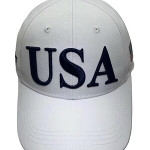 May include: White baseball cap with the word "USA" embroidered in navy blue across the front. The cap has a curved brim and a button on top. A small flag design is visible on the side.