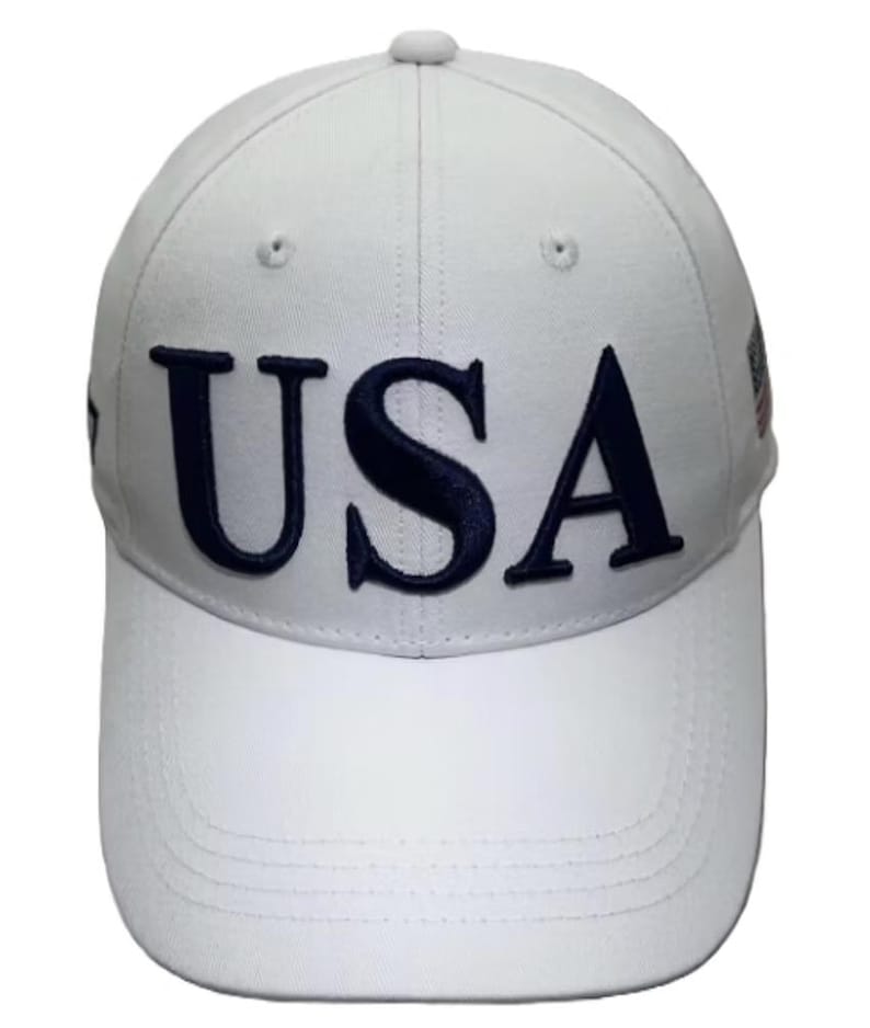 May include: White baseball cap with the word "USA" embroidered in navy blue across the front. The cap has a curved brim and a button on top. A small flag design is visible on the side.