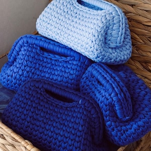 Peut inclure: Quatre sacs à main crochetés à la main dans des tons de bleu, du clair à l'indigo profond, sont disposés dans un panier en osier. Chaque sac a une forme arrondie avec une poignée. Les sacs sont faits de fil épais et texturé.
