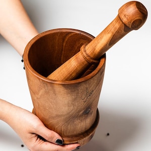 Peut inclure: Un ensemble de mortier et pilon en bois. Le mortier est un bol profond avec un large bord, et le pilon est de forme cylindrique arrondie. Les deux pièces sont en bois brun clair et sont tenues par des mains.