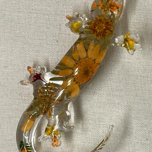 May include: A clear resin gecko with pressed flowers and leaves inside. The gecko is in a curled position with its tail curled around its body. The flowers are mostly yellow and orange, with some green leaves.
