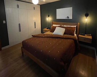 May include: A bedroom with a wooden bed frame and a brown duvet cover. The bed is against a dark green wall with a white framed picture above it. There are two bedside tables with lamps on either side of the bed.