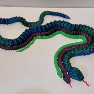 May include: Three articulated snake toys in shades of blue, green, and purple. The snakes are arranged in a winding pattern, with their heads and tails visible. The snakes have textured scales and red tongues.
