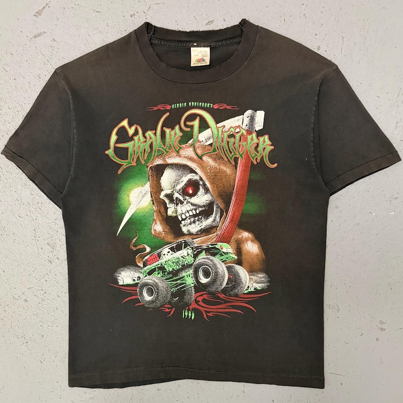 May include: Black t-shirt with a graphic of a skull in a hooded cloak with red eyes, holding a scythe. A green monster truck is below the skull. The text "Grave Digger" is above the skull.