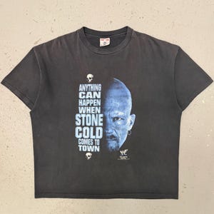 Könnte beinhalten: Ein anthrazitfarbenes Kurzarm-T-Shirt mit einer bläulichen Grafik eines Gesichts auf der rechten Seite. Der Text lautet "ANYTHING CAN HAPPEN WHEN STONE COLD COMES TO TOWN", ergänzt durch kleine Totenkopf-Symbole und ein WWF-Logo.