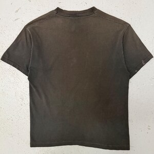 May include: A dark grey t-shirt with a crew neck and short sleeves. The garment has a distressed appearance, with signs of wear around the collar and sleeves. The fabric looks soft and worn, possibly indicating a vintage or pre-owned item.