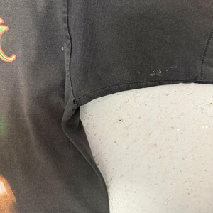 May include: Close-up of a dark grey t-shirt sleeve. The fabric shows signs of wear and tear, with small imperfections. The sleeve's edge is visible, and the shirt appears to be vintage or well-worn.