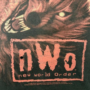 以下が含まれることがあります： 黒いTシャツ。唸るオオカミのグラフィックと、赤い長方形の中に「nWo new world Order」の文字がプリントされています。シャツはヴィンテージ風で、色が薄れており、下部には「1998 World Championship Wrestling」の文字があります。