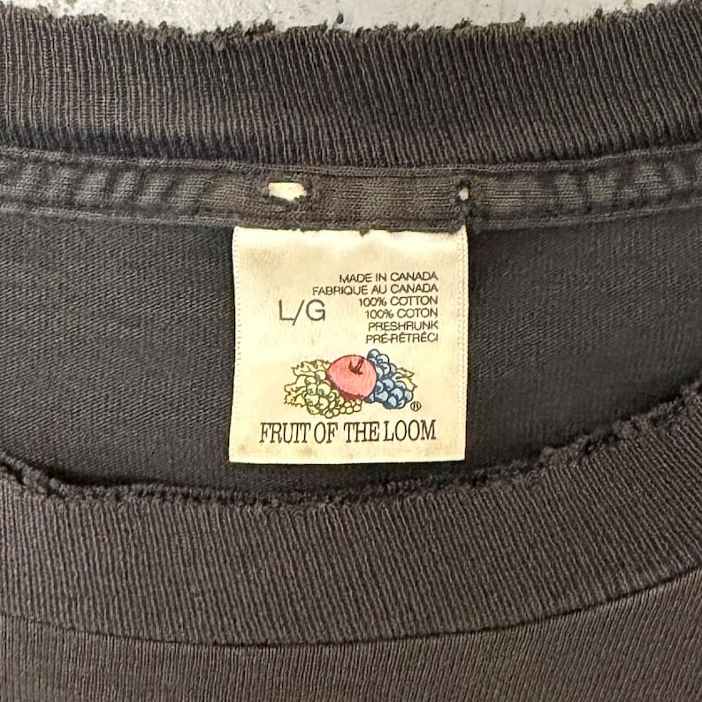 May include: Close-up of a dark grey t-shirt collar with a Fruit of the Loom label. The label reads "Made in Canada" and "100% Cotton." The label also features a graphic of an apple, grapes, and leaves. The shirt is size L/G.