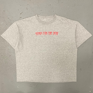 Puede incluir: Una camiseta gris jaspeada con el texto rojo "SONGS FOR THE DEAF" en el pecho. La camiseta tiene cuello redondo y mangas cortas. El fondo es una superficie gris lisa.