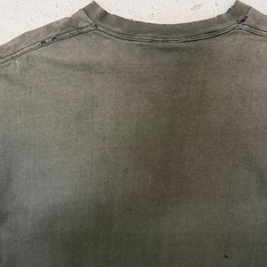 以下が含まれることがあります： 色あせたオリーブグリーンのTシャツのクローズアップ。首元が見え、縫い目に沿って小さな穴があり、使い古された、風化した外観を示しています。生地はわずかにテクスチャード加工されており、微妙な色のバリエーションがあります。