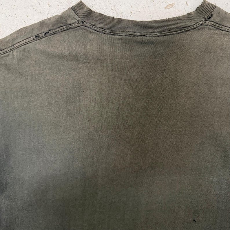 以下が含まれることがあります： 色あせたオリーブグリーンのTシャツのクローズアップ。首元が見え、縫い目に沿って小さな穴があり、使い古された、風化した外観を示しています。生地はわずかにテクスチャード加工されており、微妙な色のバリエーションがあります。