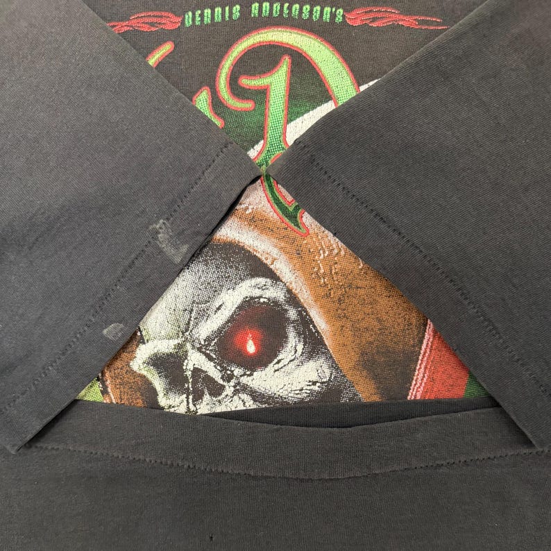 May include: Dark grey t-shirt with a skull graphic and the text "Dennis Anderson's". The design incorporates green and red details. The sleeves are visible.