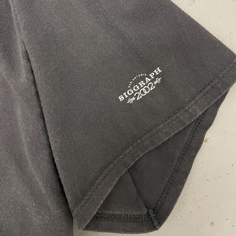 May include: Close-up of a dark gray t-shirt sleeve. The text "SIGGRAPH 2002 SAN ANTONIO" is printed in white on the sleeve. The shirt appears to be made of a soft, worn fabric.