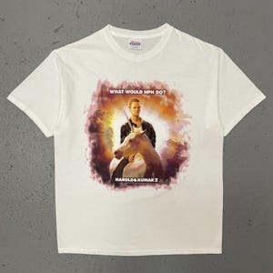 May include: White t-shirt with a graphic of a man on a white horse, text reading "WHAT WOULD NPH DO?" and "HAROLD & KUMAR 2". The design has a colourful, painterly style, with a background of orange, purple and yellow.