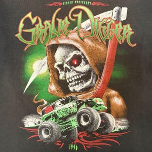May include: Black t-shirt with a graphic of a skull with red eyes in a brown hood, holding a red scythe. A green monster truck with "Grave Digger" is in front of the skull. The year "1996" is at the bottom.