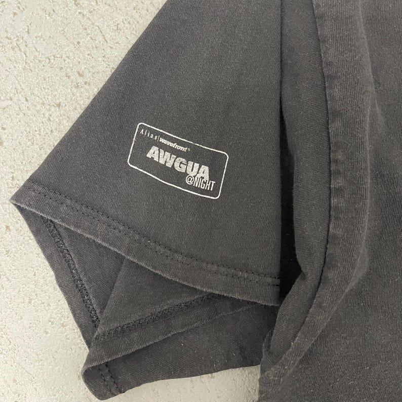 May include: Close-up of a dark gray t-shirt sleeve. A white rectangular graphic on the sleeve reads "AWGUA @NIGHT" with "Alias Wavefront" above it. The fabric has visible texture.