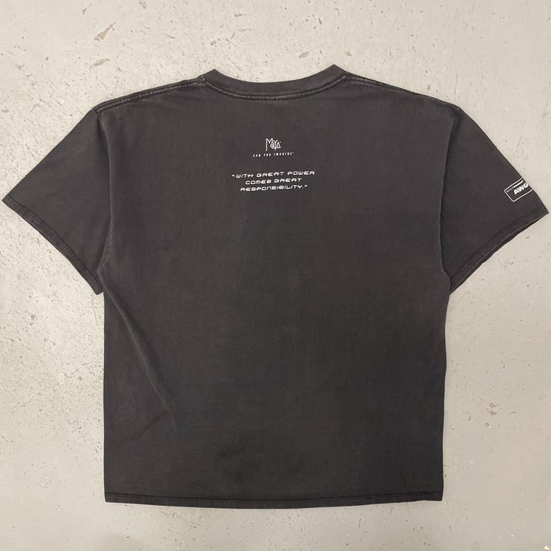 May include: Dark gray t-shirt with the text "WITH GREAT POWER COMES GREAT RESPONSIBILITY." in white. The shirt has a crew neck and short sleeves. A small logo is on the sleeve.